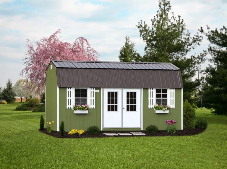 A charming green lofted barn-style shed with white double doors and decorative white shutters. The shed features flower boxes with vibrant blooms, surrounded by a well-manicured lawn and garden. A blooming pink tree and lush greenery provide a picturesque backdrop under a partly cloudy sky