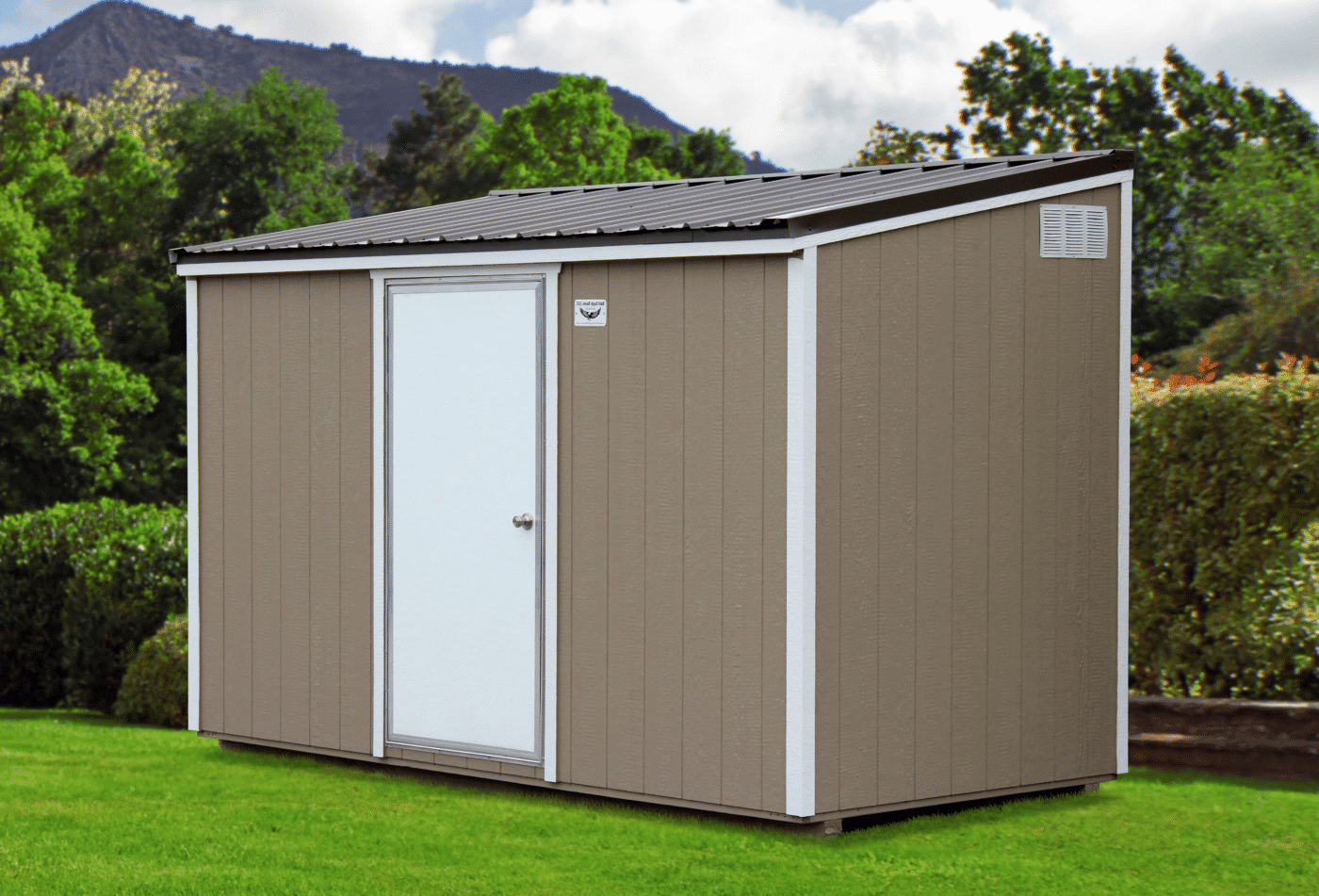 Tan utility shed with a sleek single-sloped roof and white accents, including a centered door, nestled in a manicured green lawn with lush bushes and a backdrop of forested hills and mountains