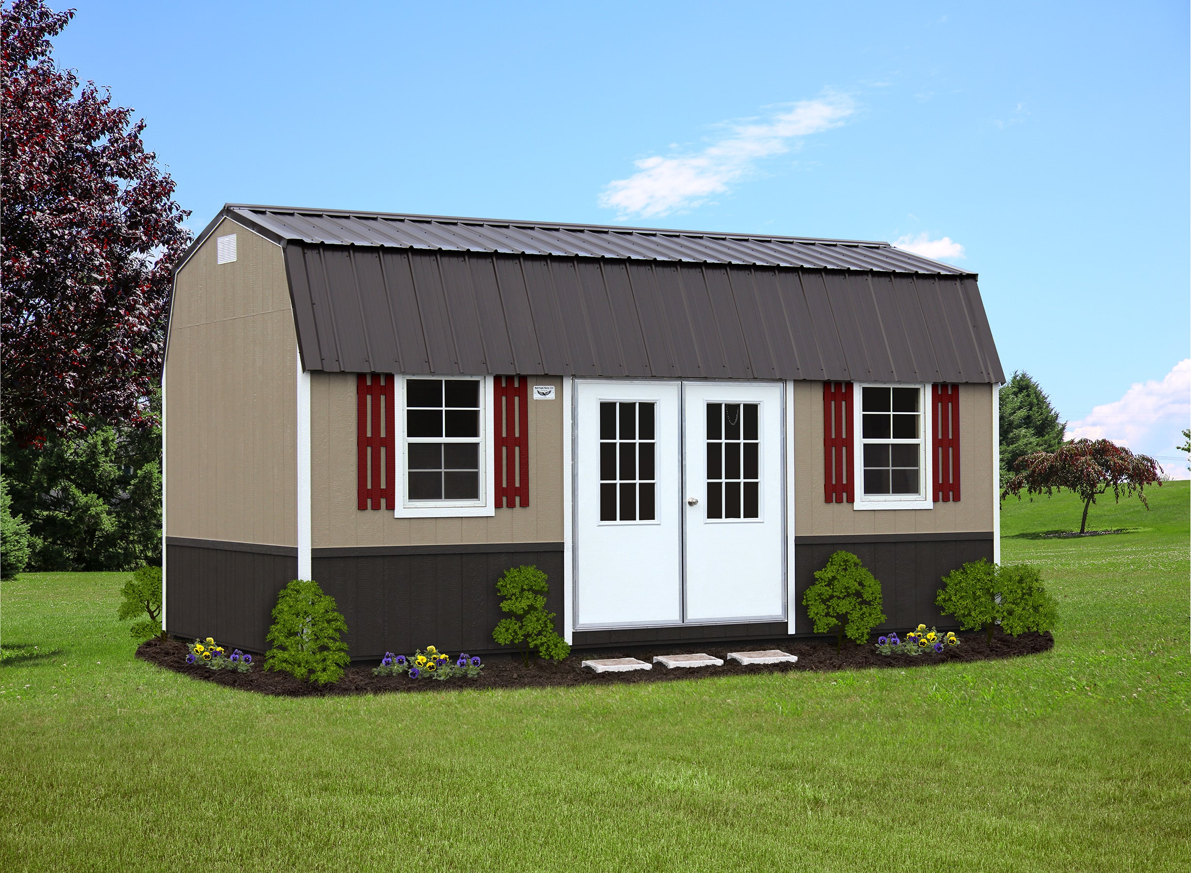A stylish side lofted barn shed featuring a beige and dark brown exterior with a metal roof. The shed includes white double doors, windows with red shutters, and beautifully landscaped surroundings with vibrant flowers and green shrubs. Set against a bright and clear sky, this shed blends functionality with aesthetic appeal