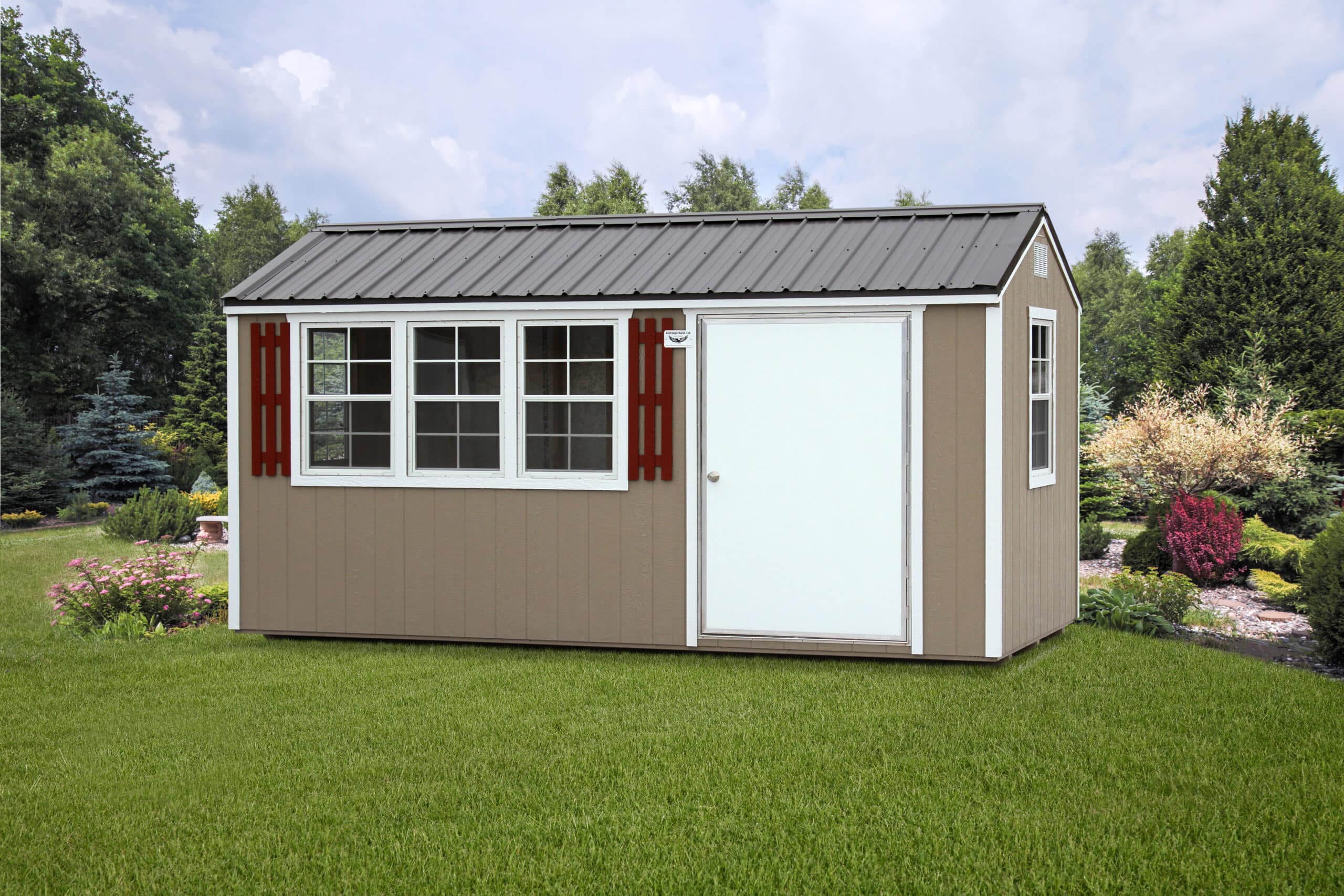 Stylish tan workshop shed featuring a white entry door, large multi-pane windows with red shutters, and a black metal roof, surrounded by a beautifully landscaped garden with vibrant greenery and colorful shrubs