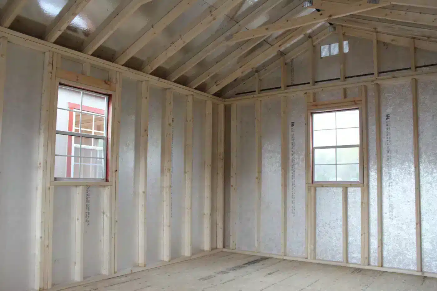 Interior view of a partially finished shed showcasing wooden framing, two windows with natural light streaming in, and insulated walls, perfect for customization into a workspace or storage area