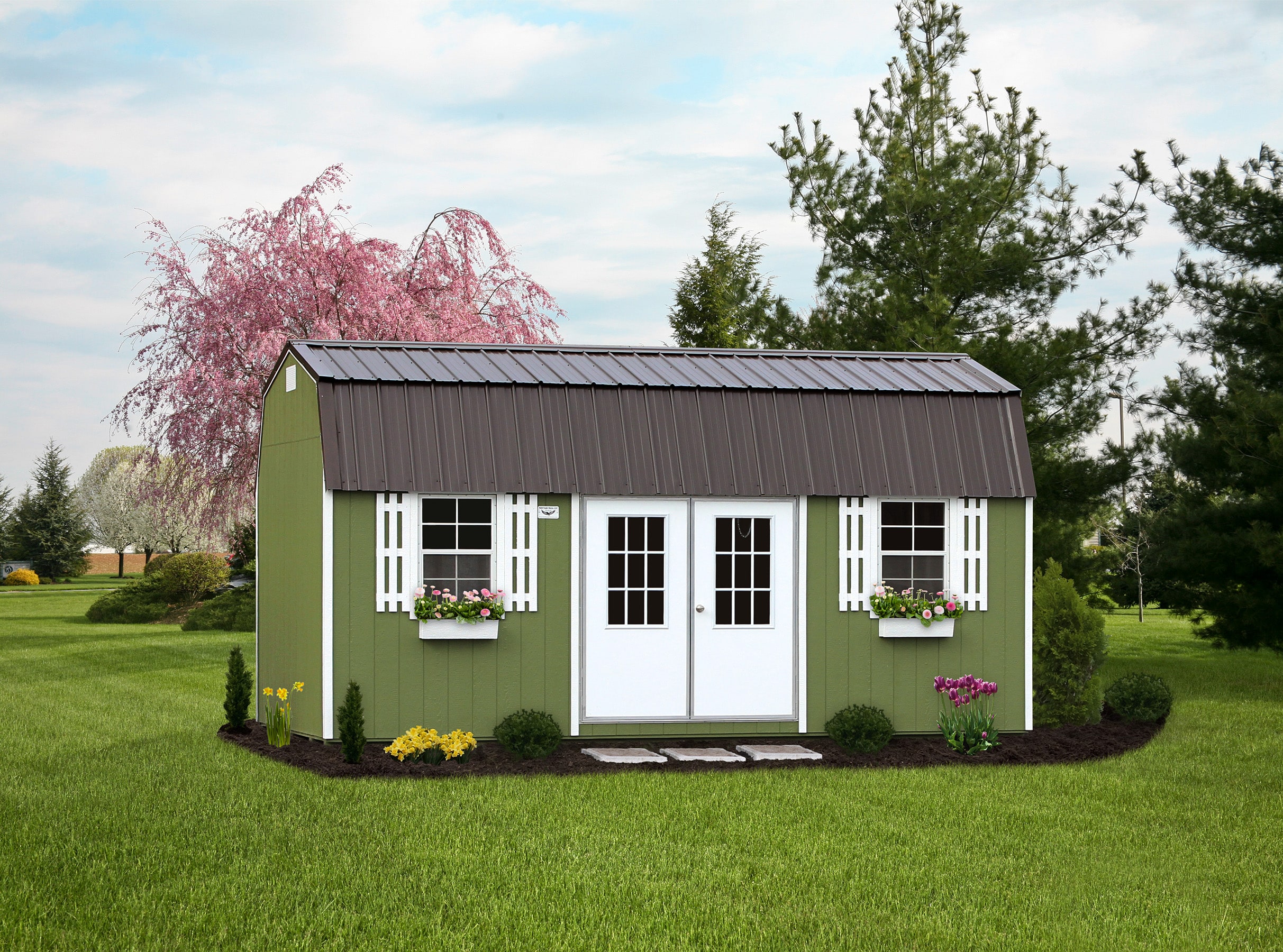 Charming green garden shed with a brown metal roof, white double doors, and decorative white shutters framing large windows adorned with flower boxes full of pink and purple blooms. Surrounded by neatly landscaped flower beds and set against a picturesque backdrop of lush trees and a blossoming pink cherry tree