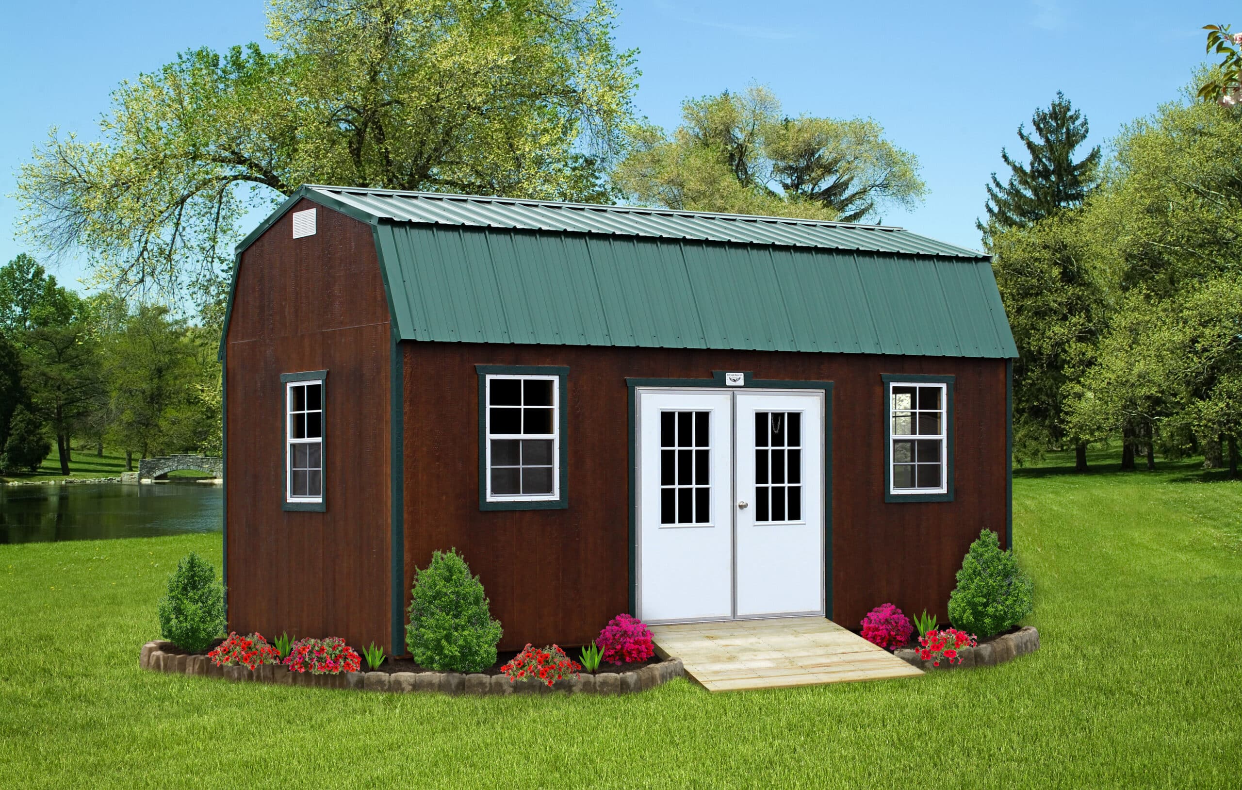 A beautiful 8x16 barn-style shed with a rustic brown exterior, accented by a green metal roof and trim, creating a classic, earthy aesthetic. The shed features double white French doors at the front, flanked by two white-framed windows that add symmetry and charm. A small wooden ramp leads to the entrance, enhancing accessibility. Surrounding the base are landscaped flower beds filled with vibrant blooms and shrubs, adding a pop of color and natural beauty. Set on a lush green lawn with a serene pond and trees in the background, the shed blends functionality with scenic elegance.