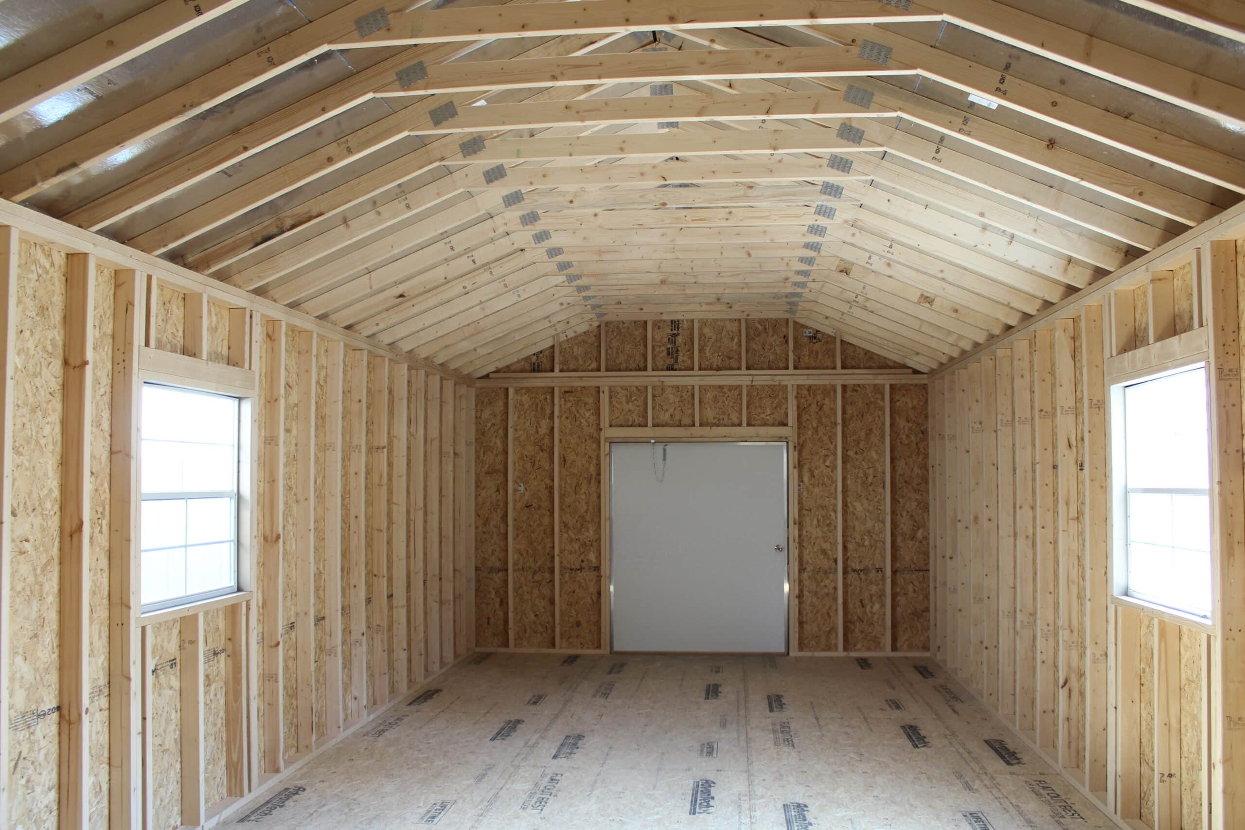 The interior of an 8x16 shed showcases an unfinished design with exposed wooden framing and OSB walls. The high, pitched roof highlights a sturdy truss system, allowing for potential lofted storage or customization. Two large windows on either side provide natural light, brightening the space and enhancing its usability. The spacious layout and unfinished state make it highly versatile, ideal for storage, a workshop, or further upgrades such as insulation or shelving. The bare plywood flooring adds to its customizable potential, offering a blank canvas for various applications.